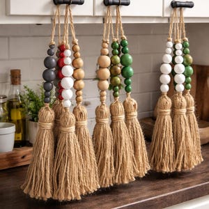 May include: Three decorative beaded tassels in different colour schemes: grey, red, and white; natural wood and green; and white and green. Each tassel has a jute fringe. The tassels hang from black hardware on a white cabinet.