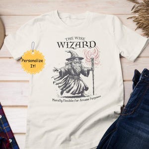 Könnte beinhalten: Ein weißes T-Shirt mit einer Schwarzweißillustration eines Zauberers, der einen Stab hält. Der Text "The Wise Wizard" befindet sich über der Illustration und "Morally Flexible for Arcane Purposes" darunter.