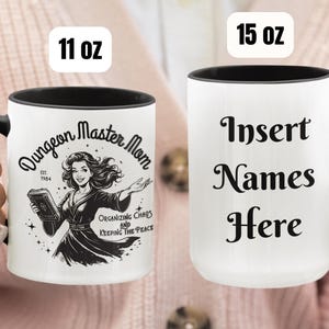 Könnte beinhalten: Zwei weiße Keramikbecher mit schwarzen Henkeln. Der Becher links zeigt eine Schwarzweißillustration einer Frau mit einem Buch und dem Text "Dungeon Master Mom Est. 1984 Organizing Chaos and Keeping the Peace". Der Becher rechts hat den Text "Insert Names Here".