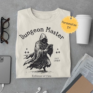 Könnte beinhalten: Ein beiges T-Shirt mit einer schwarzen Grafik einer Kapuzenfigur, die ein Buch hält. Der Text "Dungeon Master" befindet sich über der Figur, und "EST 1989" darunter. Der Text "Enforcer of Fate" befindet sich am unteren Rand der Grafik.