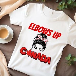 May include: White t-shirt with a red and black graphic design. The graphic features a woman with a bun in her hair and a red and white polka dot headband. The text "Elbows Up Canada" is printed in red letters.