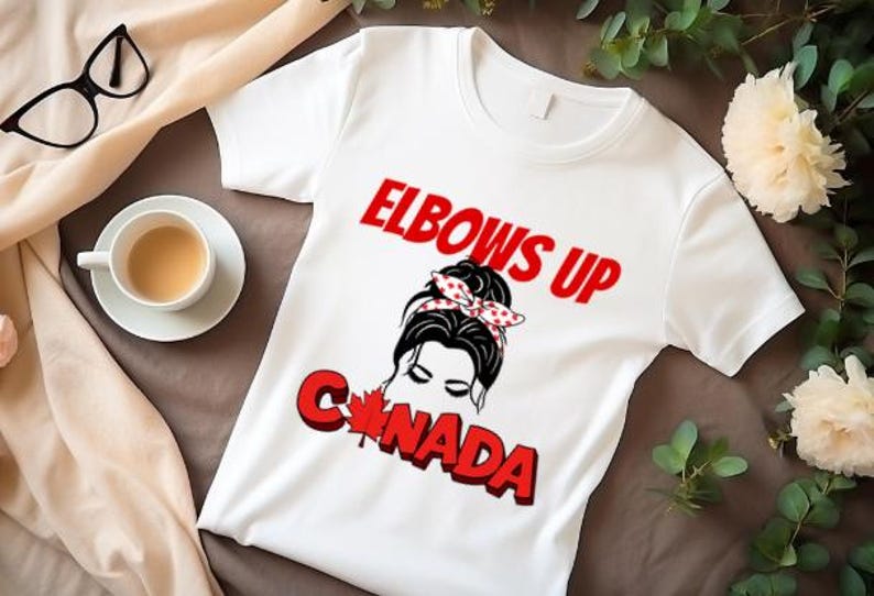 May include: White t-shirt with a red and black graphic design. The graphic features a woman with a bun in her hair and a red and white polka dot headband. The text "Elbows Up Canada" is printed in red letters.