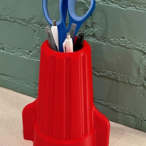 May include: A red rocket-shaped desk organizer holding blue-handled scissors, pens, and markers. The organizer has a cylindrical body with vertical grooves and a base with fins. The background is a textured, green brick wall.