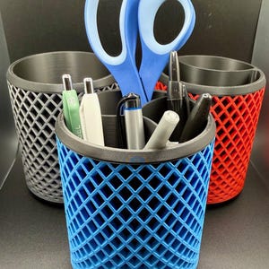 May include: Three colorful pen holders with a diamond lattice design. The holders are blue, red, and gray. A pair of blue scissors and several pens are inside the blue holder. The holders are made of plastic.