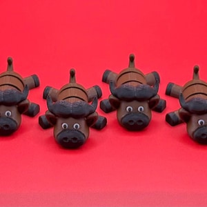 May include: Four brown and black buffalo figurines with large horns and white eyes are arranged on a red background. The buffalo are lying down, facing the viewer, and have a cartoonish appearance. The figurines are likely decorative items.