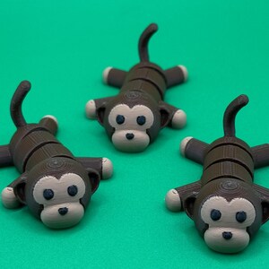 May include: Three brown and beige monkey figurines with segmented bodies and large eyes. The monkeys are lying on a green surface.