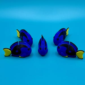 May include: Five blue tang fish figurines with black and yellow accents are arranged on a blue surface. The fish have a rounded shape with a yellow tail fin. The figurines are facing different directions.