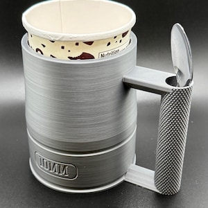 May include: A gray, 3D-printed coffee cup holder with a textured handle and base. The holder has a slot for a spoon and holds a disposable paper cup. The base of the holder has the text "10MM".