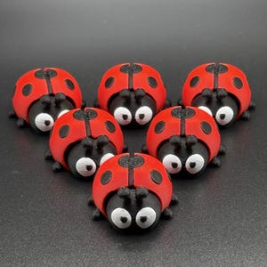 May include: A group of six 3D-printed ladybug figurines. Each ladybug has a red shell with black spots, a black head, and white eyes. The ladybugs are arranged in a circular pattern on a dark surface.