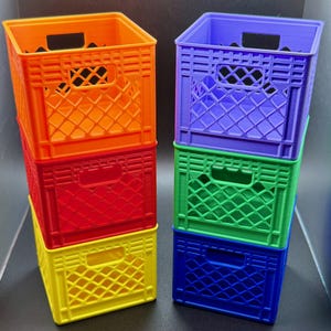 May include: A stack of four colorful miniature crates in orange, red, yellow, and purple. The crates have a rectangular shape with a lattice design on the sides and a handle cutout. The crates are stacked on top of each other.