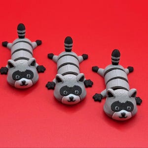 May include: Three 3D printed raccoon figurines, each with a black and white striped body and a black and white face. The raccoons are lying on their bellies with their tails curled up.