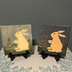 May include: Two square ceramic tiles featuring a raised relief design of a rabbit sitting in grass. The left tile has a light green glaze, while the right tile has a dark blue glaze. Both tiles depict the same rabbit figure in a pale yellow tone.
