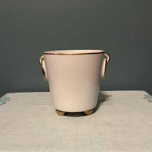 Könnte beinhalten: Ein weißer Keramik-Pflanztopf mit goldenen Verzierungen und Akzenten. Der Pflanztopf hat zwei Griffe und drei kleine Füße, alle mit goldenen Details. Der Innenraum ist weiß und die Außenseite hat eine cremefarbene Farbe.