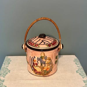 Puede incluir: Un tarro de galletas de cerámica rosa vintage con asa y tapa tejidas. La tapa presenta un borde negro y detalles florales. El exterior del tarro representa una escena con figuras con ropa de época. El asa es de mimbre tejido.