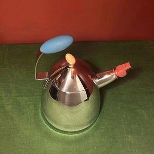 Op de afbeelding: Een theeketel van gepolijst roestvrij staal met een conisch lichaam, voorzien van een lichtblauw ovaal handvat, een kleine beige knop op het deksel en een koraalkleurig fluitje aan de tuit. Het metalen oppervlak is reflecterend en glad van textuur.