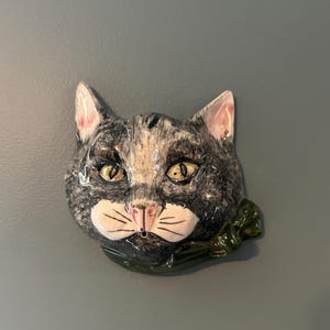 Könnte beinhalten: Eine Wandplakette aus Keramik in Form eines Katzenkopfes mit grau-weißem Fell, gelben Augen und einer rosa Nase. Die Katze trägt eine dunkelgrüne Schleife um den Hals. Das Stück hat ein kleines Loch im Mundbereich und eine glänzende Oberfläche.