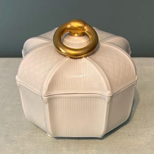 May include: White ceramic lidded trinket box with a gold ring handle. The box has a geometric shape with vertical and diamond-patterned detailing. The lid is domed, and the handle is a solid gold ring.