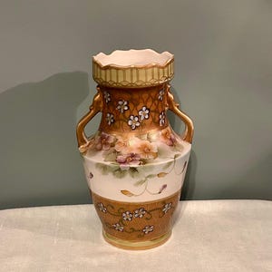 May include: A decorative vase with a cream-coloured interior and a flared rim. The vase features a brown and gold floral design with two gold handles. The body of the vase is white with painted flowers and vines.