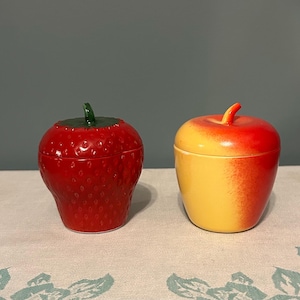 May include: Two ceramic lidded containers shaped like fruit. One is a bright red strawberry with a textured surface and a green stem lid. The other is a smooth apple with a gradient of yellow and red, featuring a small red stem on the lid.