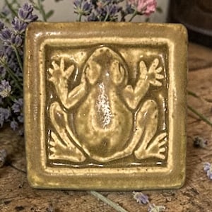 Könnte beinhalten: Quadratische Keramikfliese mit einem erhabenen Froschrelief. Der Frosch ist innerhalb eines vertieften Rahmens zentriert. Die Fliese hat eine gedämpfte olivgrüne Farbe. Der Frosch hat ausgestreckte Gliedmaßen. Die Fliese dient wahrscheinlich dekorativen Zwecken.