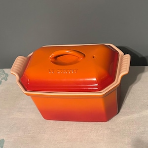 May include: A rectangular Le Creuset stoneware butter crock with a vibrant orange gradient finish. The piece features a matching lid with a central handle, integrated side handles with ribbed detailing, and a light cream interior rim.