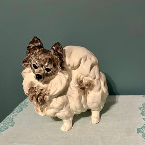 May include: A ceramic figurine of a dog, primarily white with brown accents. The dog has a fluffy appearance with detailed fur and a small, alert face. The figurine is standing on a light blue surface.