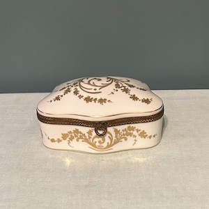 May include: A small, white porcelain trinket box with a gold floral design and a gold-colored metal trim. The box has a hinged lid and a clasp. The floral design is repeated on the lid and the base of the box. The box is sitting on a white surface.