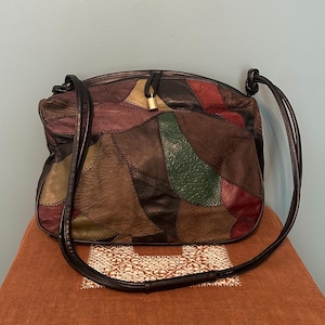 May include: A patchwork shoulder bag constructed from various leather pieces in shades of brown, black, green, and burgundy. The bag features a top zipper closure with a small gold-toned padlock charm attached to the center and two thin black straps.