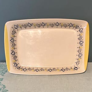 以下が含まれることがあります： クリーム色の中心と黄色の縁を持つ長方形のサービングプラッター。縁は、青と黄色の様式化された花の要素の繰り返し模様で装飾されています。ヴィンテージ品。