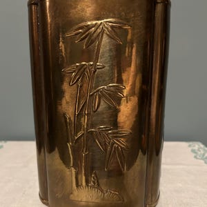 Puede incluir: Un recipiente rectangular de color bronce con un diseño de bambú. El recipiente tiene un acabado brillante y un relieve detallado de tallos y hojas de bambú. El diseño está centrado en la parte delantera, añadiendo un toque de decoración inspirada en la naturaleza.