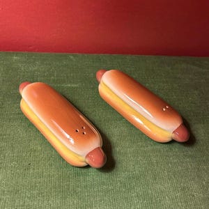 May include: A pair of ceramic salt and pepper shakers shaped like hot dogs. Each shaker features a brown sausage nestled in a yellow bun with a glossy finish. One shaker has three small holes on the top, while the other has two small holes.