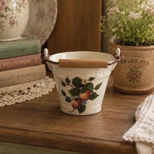 May include: A small, white ceramic bucket with a wooden handle and metal accents. The bucket is decorated with a strawberry and leaf design. Other items in the image include books, a vase, and a potted plant.