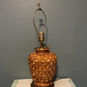 Puede incluir: Una lámpara de mesa decorativa con una base tejida y texturizada y un cuello y parte superior de metal color latón. La lámpara tiene una base marrón en forma de piña y una base de madera redonda y marrón oscuro. La lámpara mide unos 51 cm de alto.