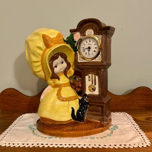 May include: A ceramic figurine featuring a brown grandfather clock with a gold-rimmed face and black hands, standing next to a figure wearing a large yellow bonnet and dress. A small black cat sits at the base, and the clock face reads Casey.