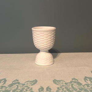 May include: A white ceramic egg cup with a woven basket design. The egg cup has a round bowl and a pedestal base. The egg cup is sitting on a light-coloured surface.