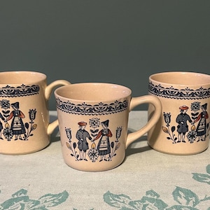 May include: Three beige ceramic mugs with handles, each adorned with a blue and white folk art design. The mugs feature a decorative border and a central image of figures in traditional attire. The mugs are displayed on a patterned surface.