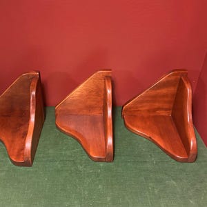 May include: Three identical corner shelves crafted from polished medium-brown wood. Each shelf features a triangular base with a curved front edge and two vertical back panels that meet at a right angle to fit securely into a wall corner.