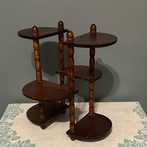 May include: A dark brown, wooden plant stand with multiple semi-circular shelves. The stand has a tiered design with three levels, supported by turned wooden posts. Each shelf is topped with a rounded finial. The stand is placed on a floral patterned tablecloth.