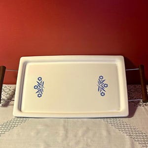 May include: A rectangular white serving tray featuring a smooth surface with two symmetrical blue floral patterns. The tray includes two dark brown wooden handles attached to the sides by thin metal rods, creating a functional serving piece.