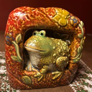 Könnte beinhalten: Eine Keramikskulptur mit einem grünen, texturierten Frosch, der in einem rechteckigen, rotbraunen Rahmen sitzt. Der Rahmen ist mit erhabenen, geformten gelben und blauen Blumen sowie grünen Blättern verziert, alles mit einer glänzenden, gesprenkelten Glasur.