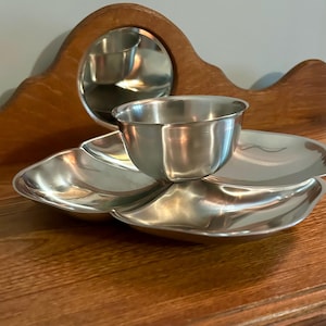May include: A silver-colored serving dish with a central bowl and three petal-shaped compartments. The dish sits on a wooden surface, with a wooden mirror in the background. The bowl and compartments reflect the light.