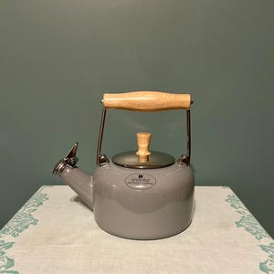 May include: A gray Chantal tea kettle with a wooden handle and lid knob. The kettle has a silver spout and a silver band around the base of the handle. The kettle is sitting on a white tablecloth with a floral pattern.