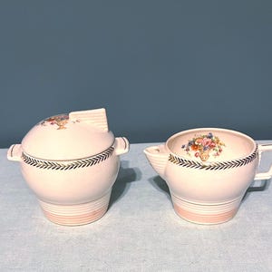 May include: A vintage cream and sugar set. The set includes a lidded sugar bowl and a creamer. Both pieces are white with a pink band at the base, a black and gold leaf design, and a floral motif. The creamer has a handle and a spout.
