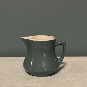 May include: A small, teal-colored ceramic creamer with a white interior rim and a curved handle. The creamer has a rounded body and a spout for easy pouring. It sits on a light-colored surface.