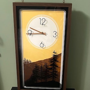May include: A rectangular wall clock with a dark brown wood-grain frame. The clock face features a yellow gradient background with a black silhouette of a forest at the bottom. It has white numbers 12, 3, 6, and 9, and thin brown clock hands.