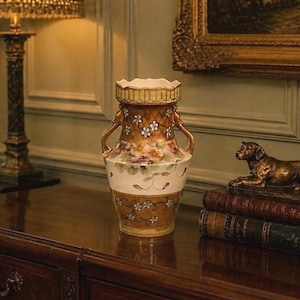 Op de afbeelding: Een decoratieve keramische vaas met bloemen- en geometrische patronen. De vaas heeft een crèmekleurige basis en een bruine bovenkant met handvatten. Een bronzen hondenbeeld staat op een stapel boeken in de buurt.