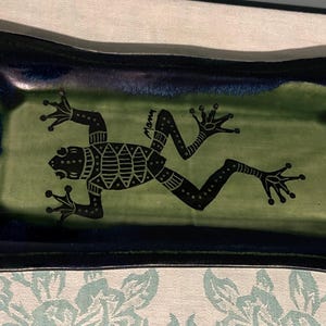 May include: A ceramic serving tray with a dark blue rim and a green interior. A black frog design is centered on the tray. The frog has a detailed pattern and the word "Mony" is written on the tray.