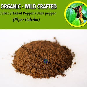 Op de afbeelding: Een close-up van een hoop gemalen cubebpeper, ook bekend als staartpeper of Java-peper. De peper is donkerbruin van kleur en heeft een grove textuur. De tekst "ORGANIC - WILD CRAFTED Cubeb/Tailed Pepper / Java pepper (Piper Cubeba)" is zichtbaar bovenaan de afbeelding.