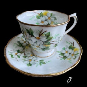 May include: White teacup and saucer set with a floral design. The teacup and saucer are white with a gold trim and feature a pattern of white flowers with green leaves and yellow accents. The set is on a black background.
