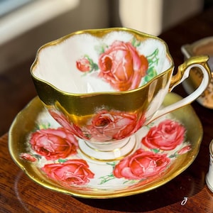 May include: A white porcelain teacup and saucer set featuring a vibrant red rose pattern with green leaves. The teacup has a scalloped rim and a handle accented with gold trim. The saucer also features a scalloped edge with a wide gold border.